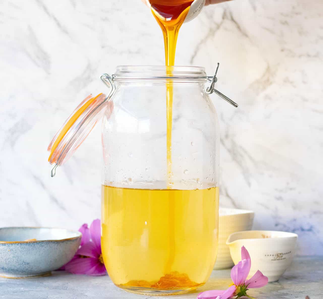A large jar filled with jun tea sits on a bench. Honey is being poured in, which can be seen settling on the bottom.