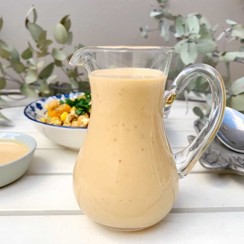 Close-up of a small jug filled with Miso Tahini Dressing sitting on a white table in front of a salad.