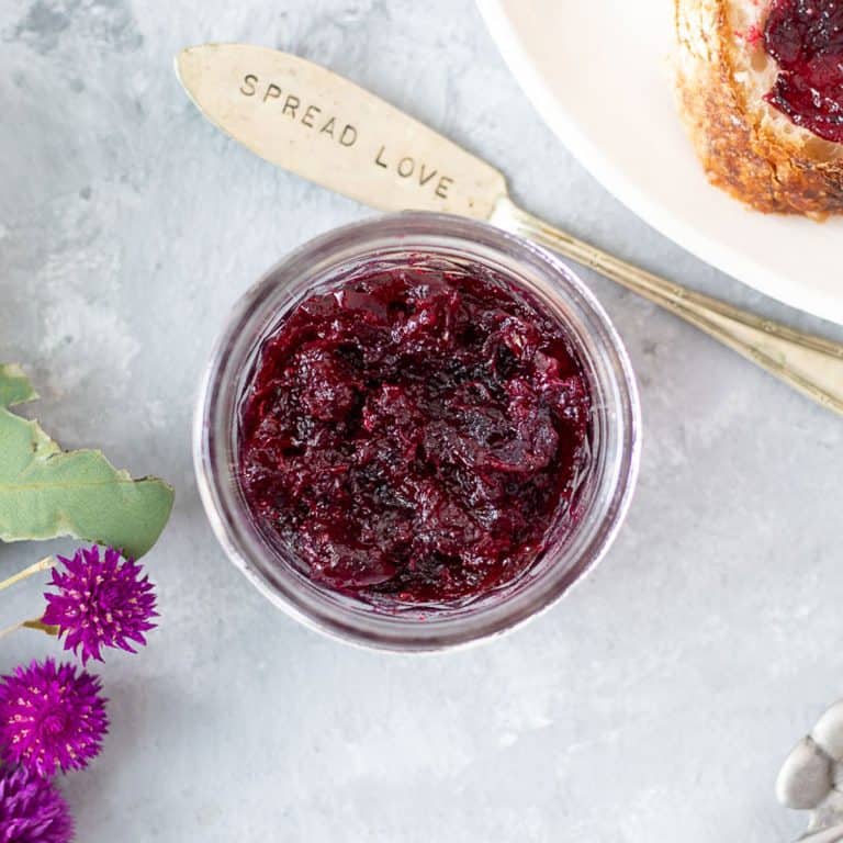 Looking down over a small jar filled with ruby red-coloured rosella jam. A knife with spread love on it sits beside the jar.