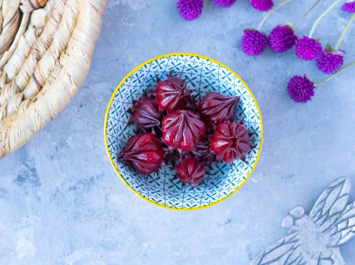 A small patterned bowl with a yellow rim sits on a grey table and is filled with ruby red-coloured Rosella fruit