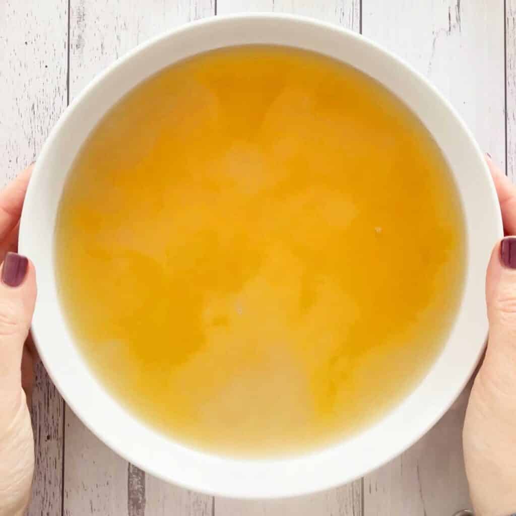 Two white hands are holding the edges of a bowl of golden chicken stock.