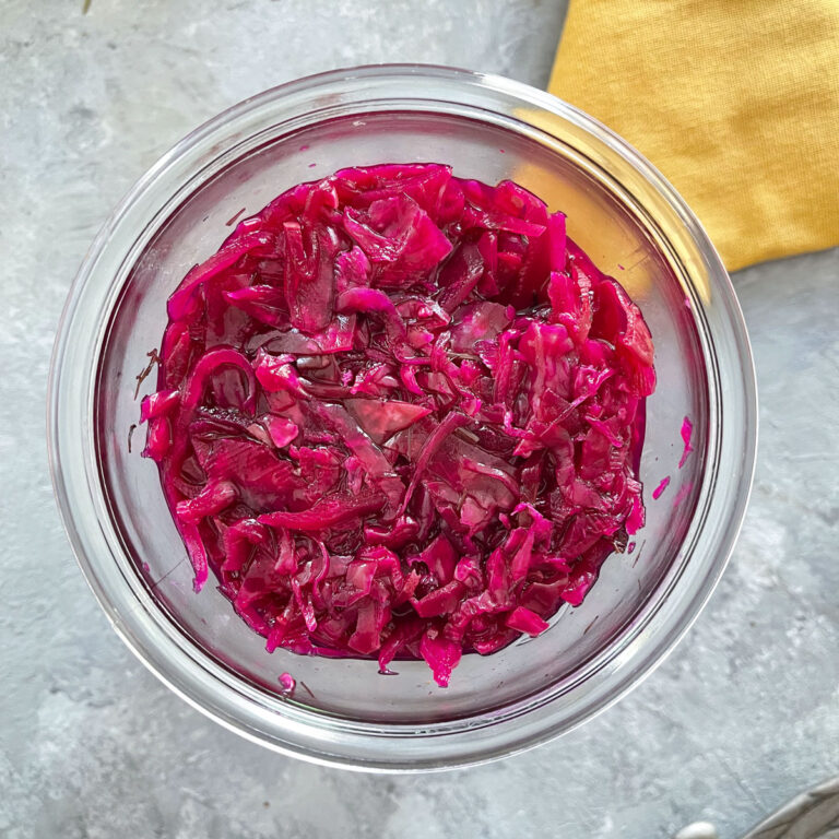 A square image showing the name of the recipe along with a square photo looking into a jar of red cabbage sauerkraut. It is sitting on a grey table with a yellow napkin.