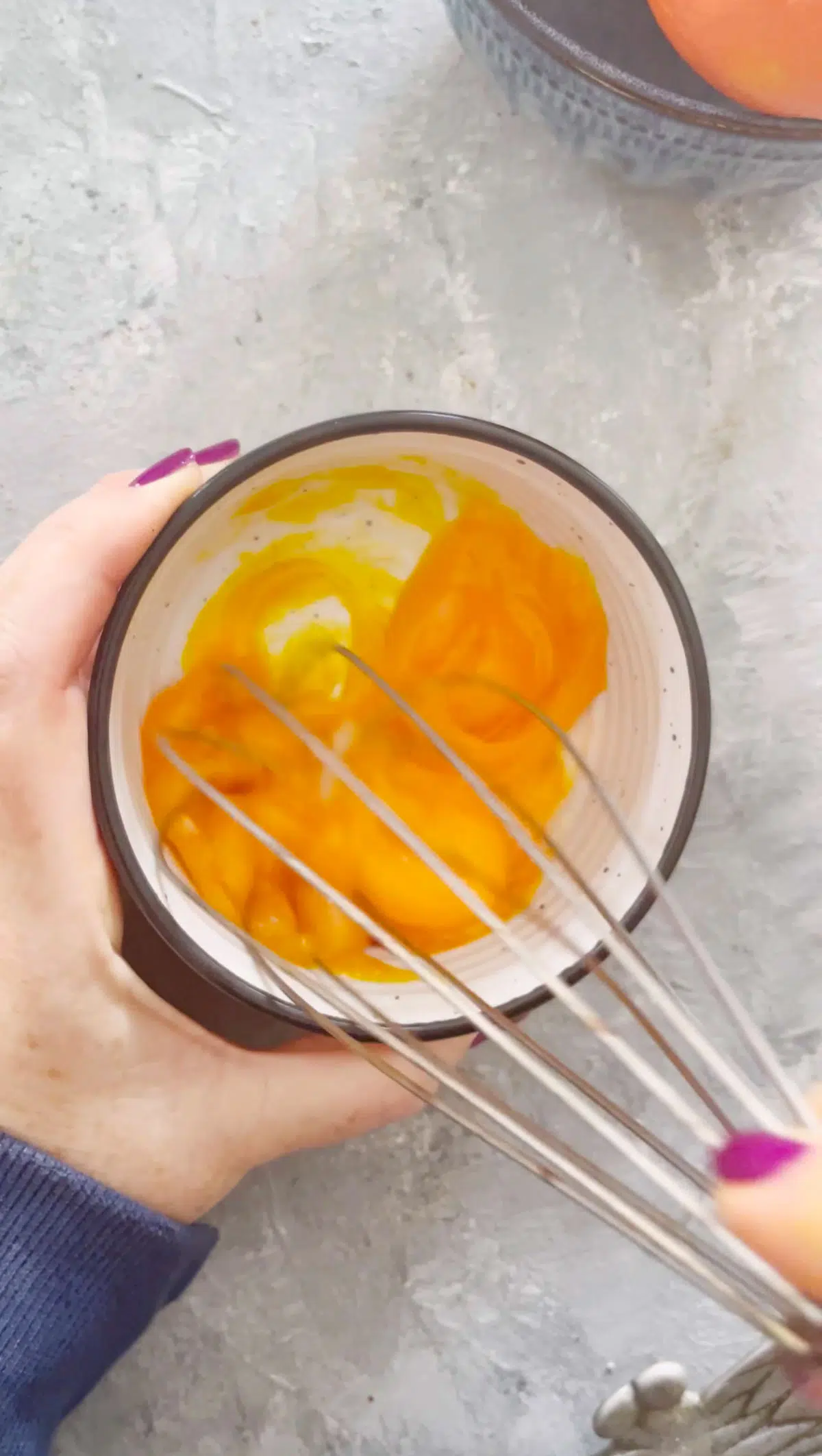 A hand is holding a small white bowl that contains 4 egg yolks. A whisk has started to whisk the yolks together.