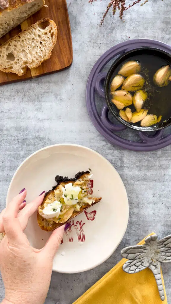 In the centre of the image is a white plate with red butterflies on it and a hand hovers above it holding a piece of toast that's been spread with garlic confit. On top of that is some mozzarella and a sprinkle of thyme. Beneath the plate on the concrete table is a yellow napkin and an ornamental dragonfly. Slightly above the plate on the table is a purple trivet underneath a small frying pan that contains the garlic confit. And well above the plate on the table to the left is a chopping board with a loaf and slices of sourdough.
