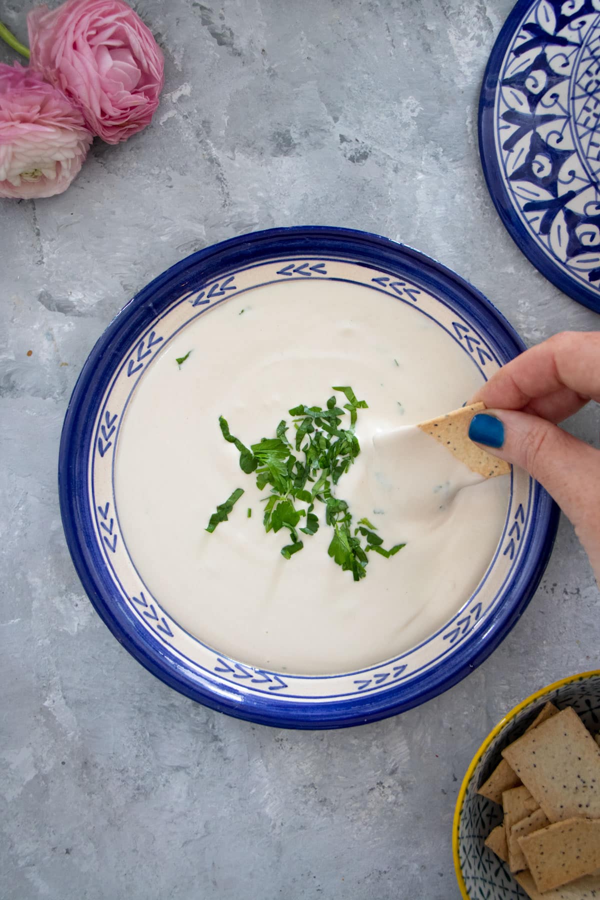 A white hand is dipping a rectangular cracker into a blue-rimmed bowl full of lemon tahini sauce.