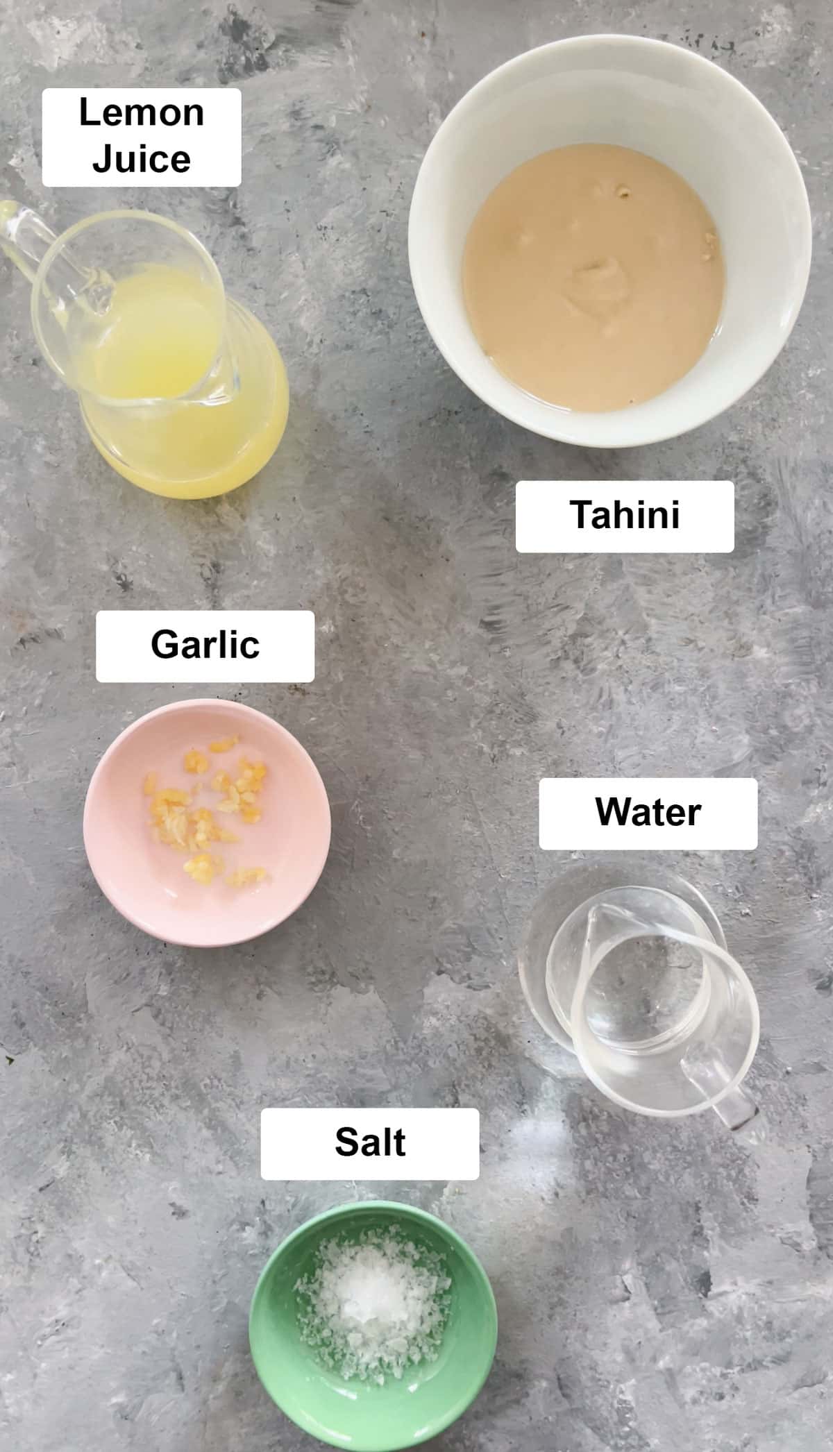 All the ingredients needed to make lemon tahini sauce are spread out across a grey table in small bowls.