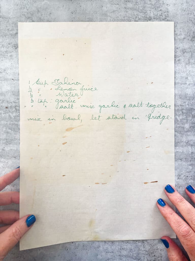 An aged piece of paper is behind held by two hands with dark blue nail polish. There is a recipe for tahina on the page.