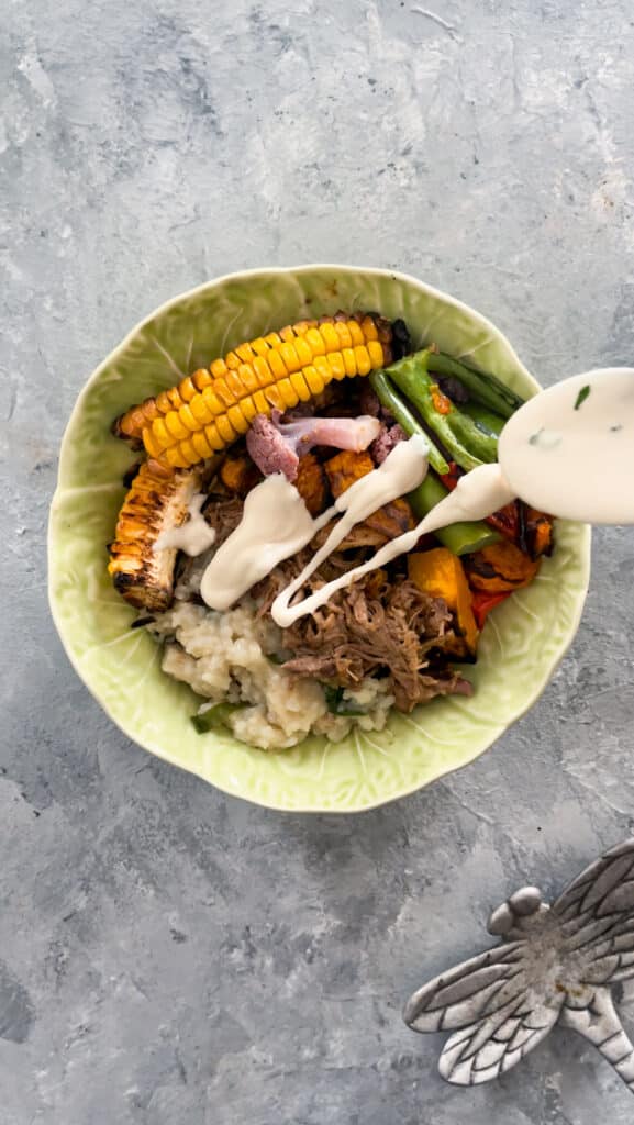 A green bowl in the shape of a cabbage is in the centre of a grey table. It is filled with roasted meat, rice, roasted vegetables and a hand holding a spoon is drizzling over some tahini sauce. To the bottom right of the image is a decorative silver dragonfly.