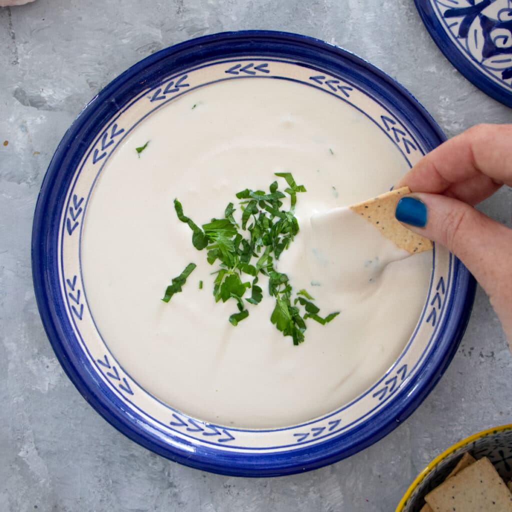A white hand is dipping a cracker into a bowl of tahini dip with shredded parsley on top.