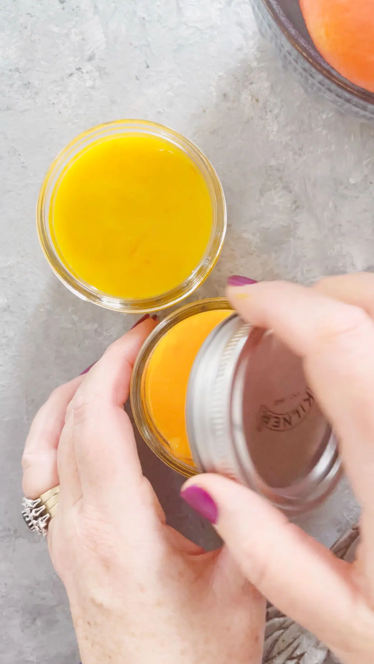 A white hand is screwing a silver lid onto a small jar of bright orange tangelo curd.