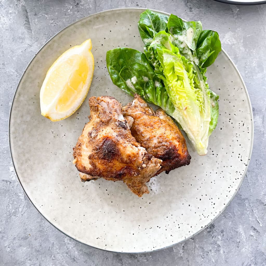 This is a square image of a stone coloured plate is in the middle of the image sitting on a grey table. On the plate are two pieces of cooked chicken with a dollop of yoghurt and a wedge of lettuce to the right on the plate, along with a wedge of lemon to the left.