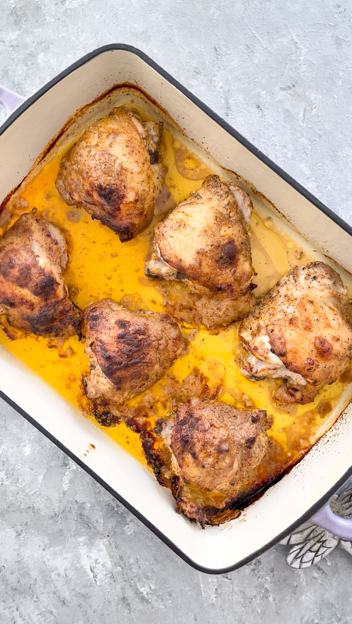 A deep baking dish is sitting on a grey table. Inside is the cooked chicken swimming in gold-coloured pan juices.