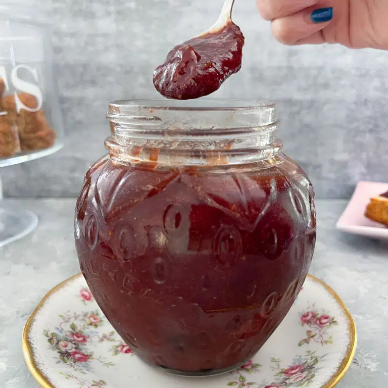 This is a square image showing a jar shaped like a strawberry is filled with rhubarb and strawberry jam. The jar is sitting on a white saucer with a floral rim. A white hand with blue nail polish is holding a teaspoon of the jam just above the jar. Behind the jar on the left is a glass cake holder filled with rustic scones. And to the right is a small plate with a scone topped with jam and cream.