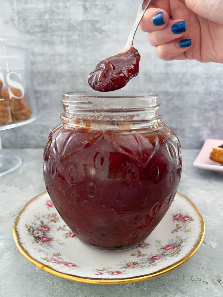 A jar shaped like a strawberry is filled with rhubarb and strawberry jam. The jar is sitting on a white saucer with a floral rim. A white hand with blue nail polish is holding a teaspoon of the jam just above the jar. Behind the jar on the left is a glass cake holder filled with rustic scones. And to the right is a small plate with a scone topped with jam and cream.