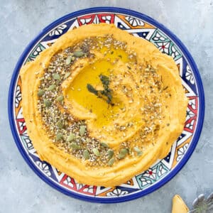This is a square image of a colourful Moroccan plate sitting on a grey table. Covering most of the plate is bright orange pumpkin hummus drizzled generously with olive oil and sprinkled with dukkha and pumpkin seeds. Some cooked thyme sits decoratively in the middle of the oil. In the bottom right hand corner below the plate, you can just make out the top of a silver ornamental dragonfly sitting on top of a yellow napkin.