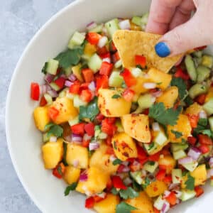 This is a square image that is close up on the bowl of mango and cucumber salad. A white hand with blue nail polish is holding a corn chip and dipping it into the salad.