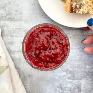 A small jar of strawberry and rose jam is sitting on a grey table. Above it and to the right is a small white plate with a small piece of tart on it that contains the jam. The plate is being held by a white hand with blue nail polish. To the left of the jam jar is a white napkin.
