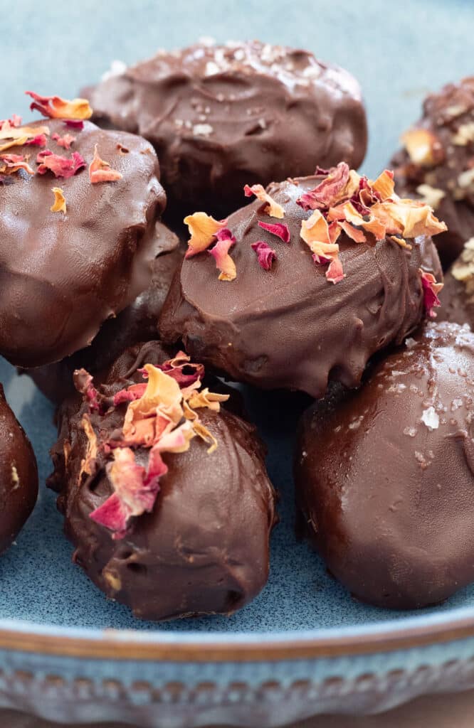 Close up of chocolate covered dates sitting in a blue bowl topped with rose petals, crushed nuts, and salt.