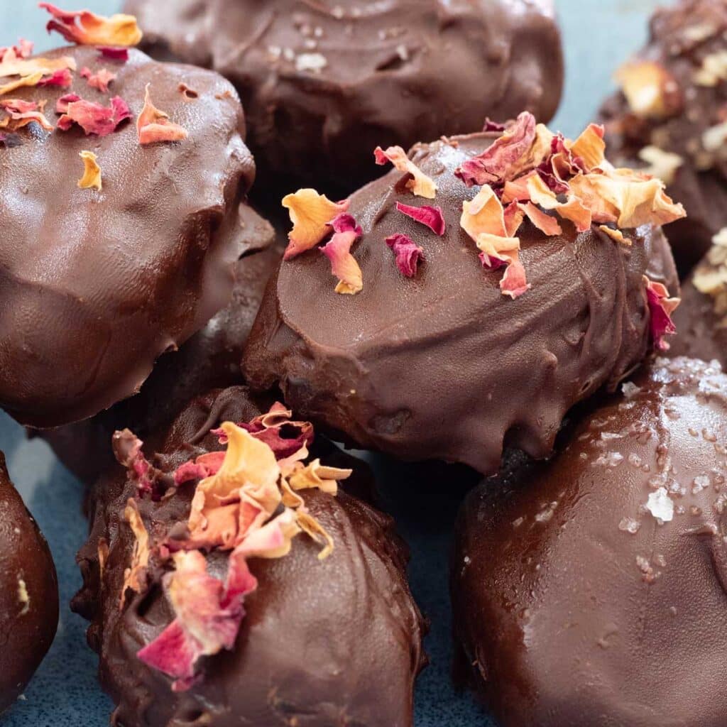 A square image showing a close up of chocolate covered dates sitting in a blue bowl topped with rose petals, crushed nuts, and salt.