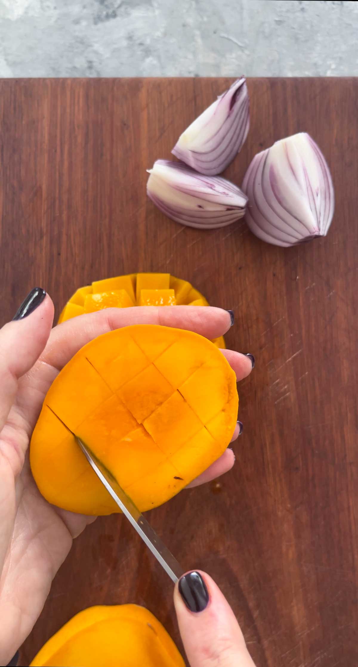 A white hand is holding a mango cheek and cutting the flesh into a crisscross with a knife.