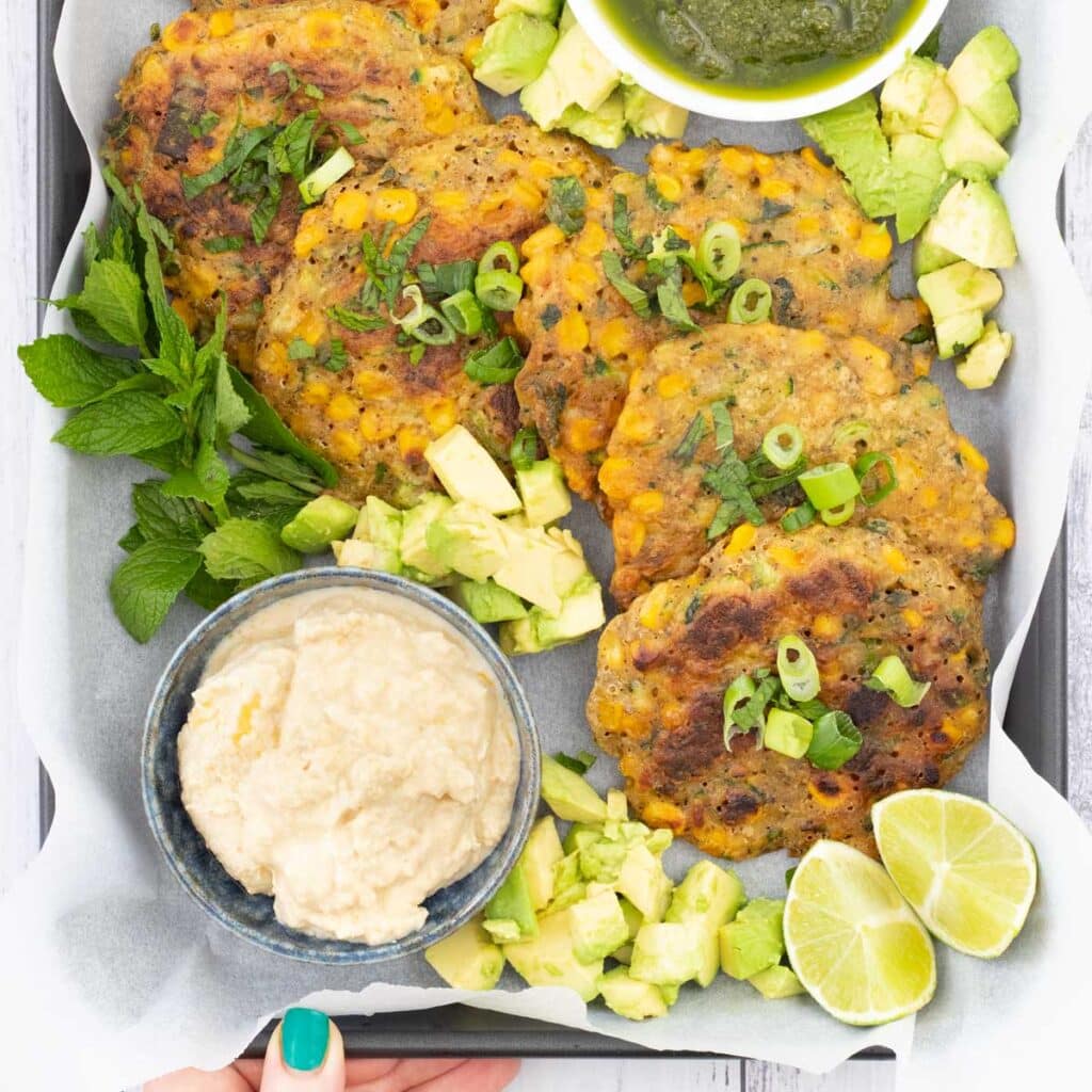 This is a square image showing a close up of a tray lined with baking paper has been topped with zucchini fritters, chopped avocado, a small bowl of pesto and one of hummus.