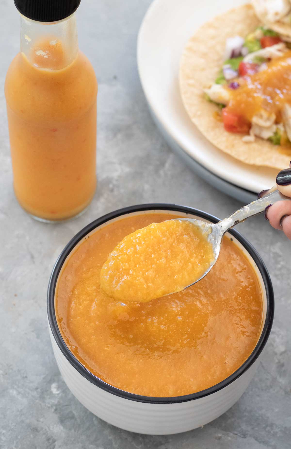 A white bowl with a black rim has been filled with bright orange mango chilli sauce. A white hand has dipped a spoon into the sauce and is holding it slightly above the bowl. The bowl is sitting on a grey table and behind it is a bottle also containing the mango chilli sauce, and a plate of tacos. 