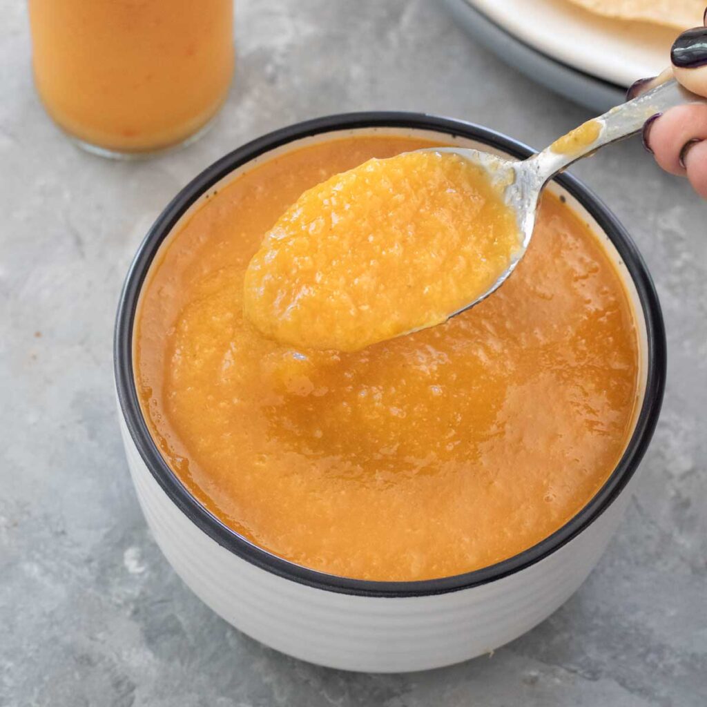 A white hand is holding a spoonful of mango chilli sauce above a white bowl with more sauce in it.