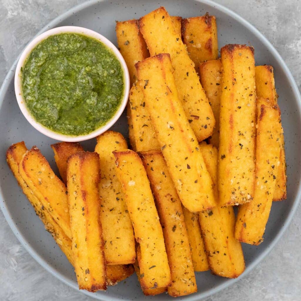 Close up of a grey/blue bowl filled with golden polenta chips and a small bowl of pesto.