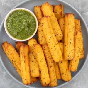 Close up of a grey/blue bowl filled with golden polenta chips and a small bowl of pesto.