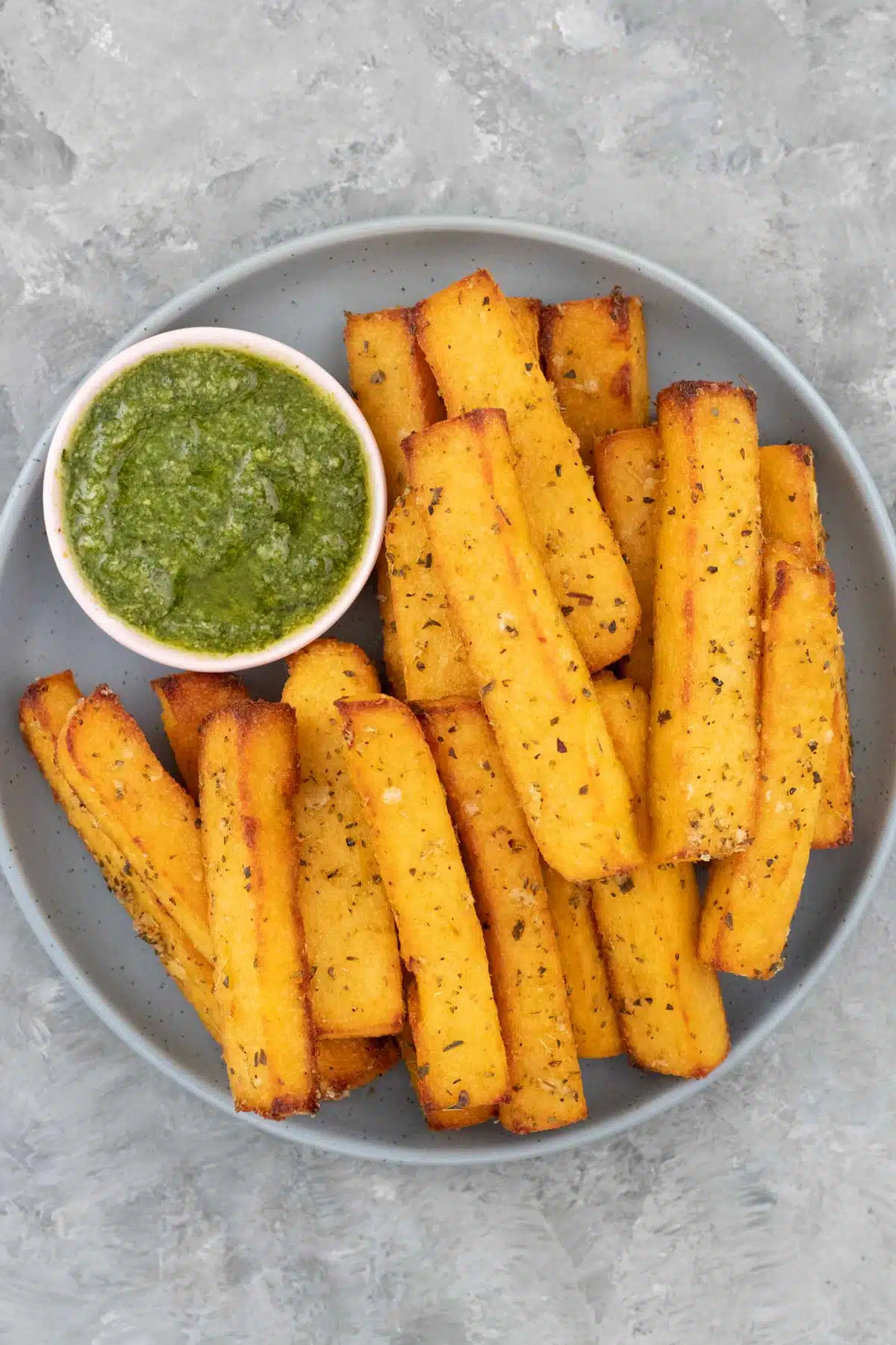 A grey/blue bowl filled with polenta chips and a small bowl of pesto is sitting on a grey table.
