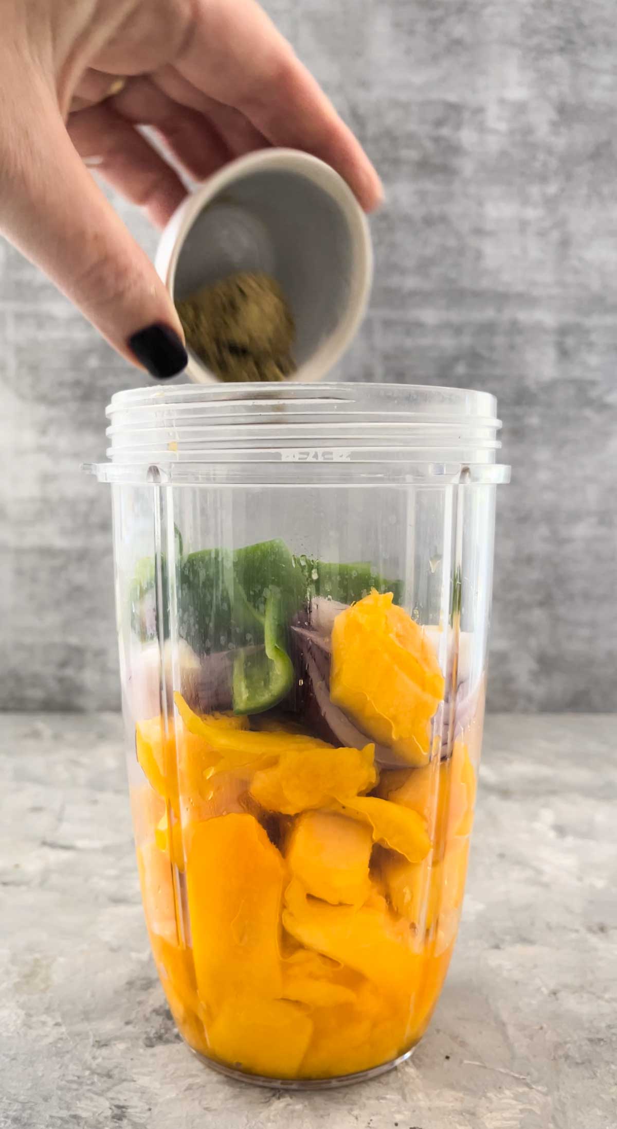 All the ingredients needed to make mango chillis sauce have been placed into a blender jug. A white hand is adding one more.