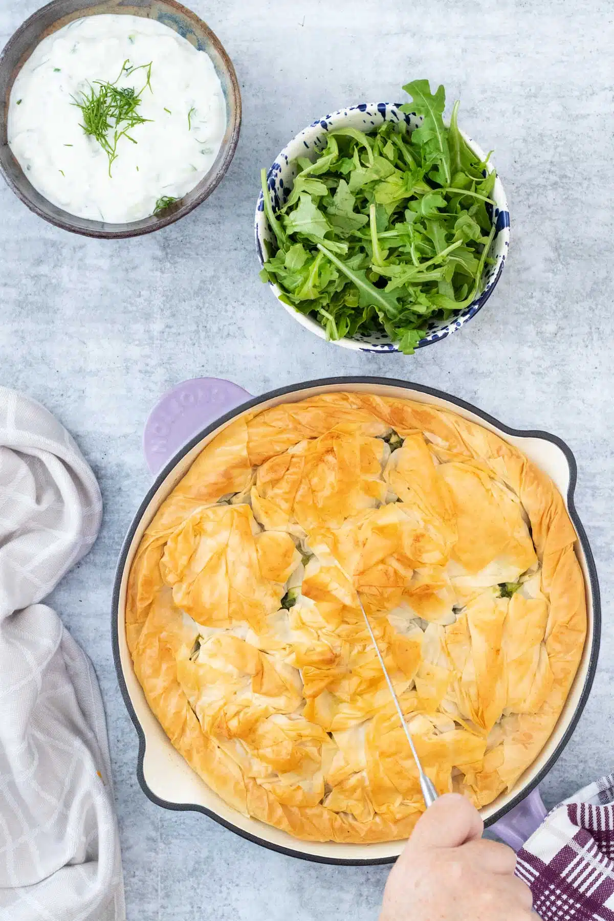 A golden brown spanakopita in a skillet is sitting on a grey table. A tea towel is to the left of it and above is a bowl of rocket and one of tzatziki. A white hand is holding a knife and cutting into it.