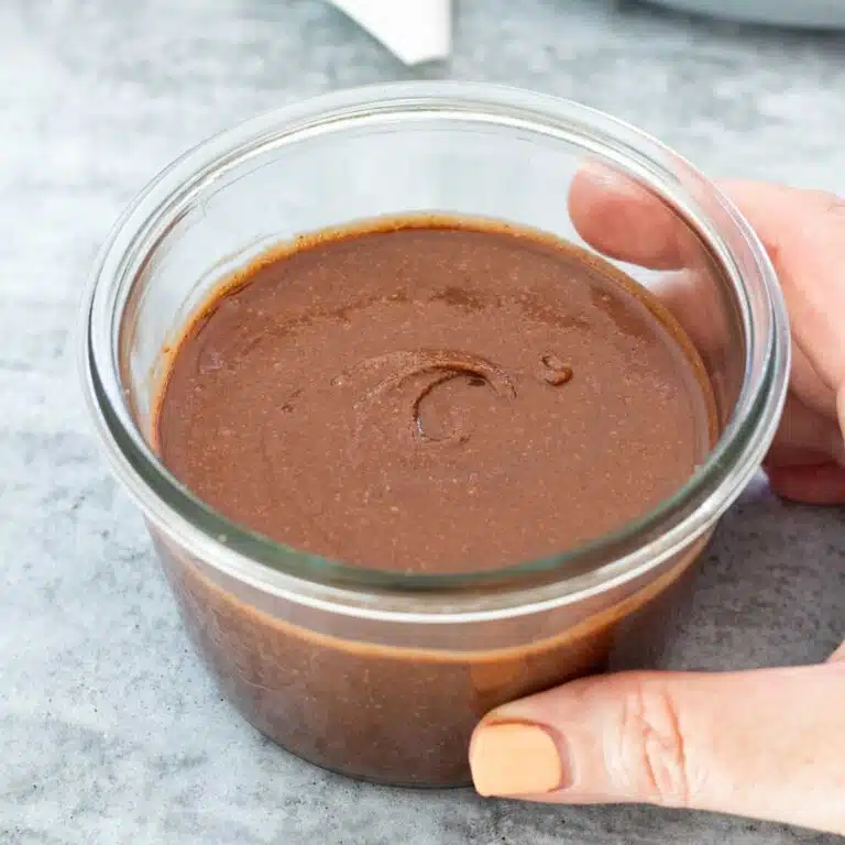 A jar of homemade chocolate spread is sitting on a grey table and being held by a white hand with orange nail polish. Square image.