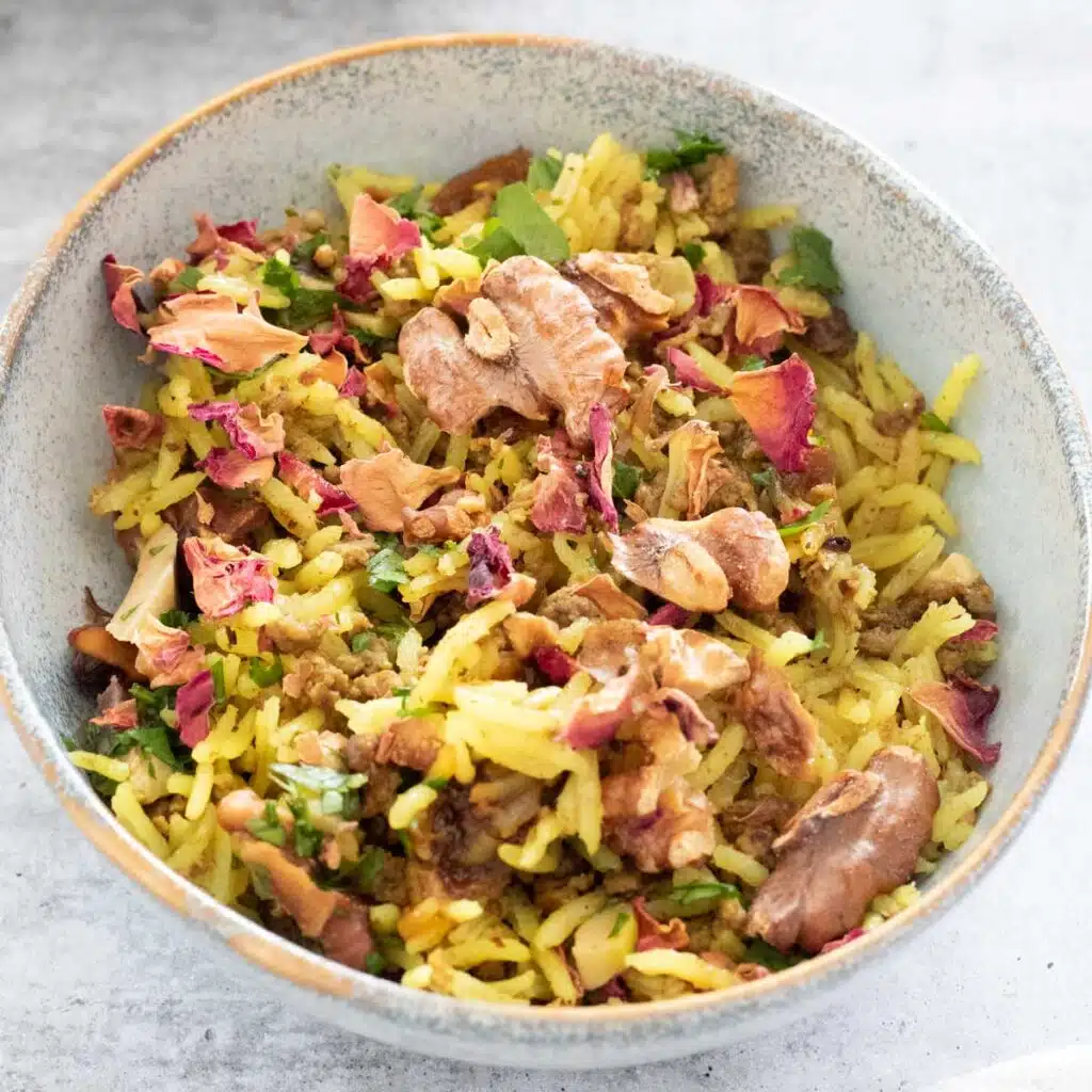 A square image of a small blue bowl filled with lamb pilaf. It's topped with chopped parsley, dried rose petals, chopped green olives and roasted walnuts.