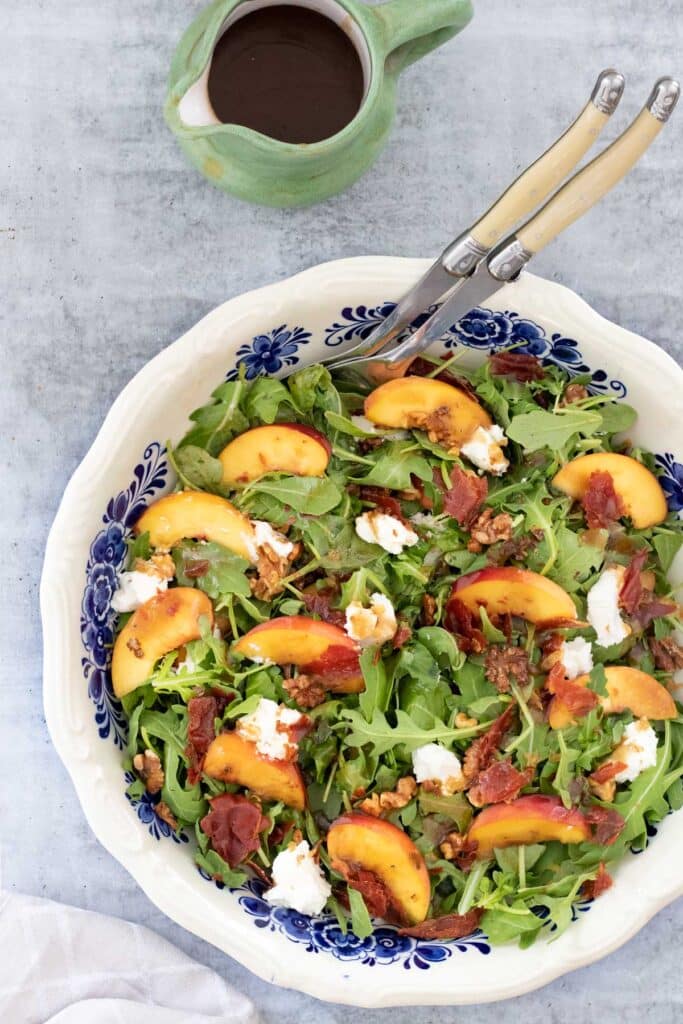 Sliced peaches, arugula, goat's cheese, walnuts and prosciutto are layered in a large white bowl with blue patterned interior