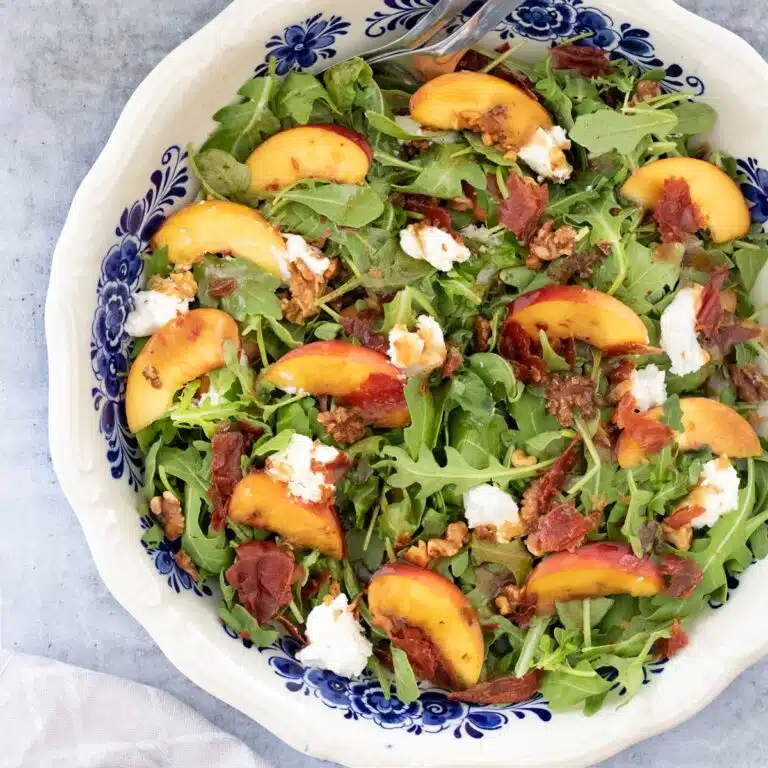 A square image showing a close up of arugula, peaches, toasted walnuts, crispy prosciutto, and soft goat's cheese tossed with dressing in a large bowl.