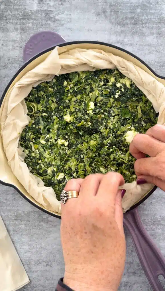Two white hands with dark pink nail polish are rolling up the overhanging edges of filo to create a crust around the edge of the skillet spanakopita.