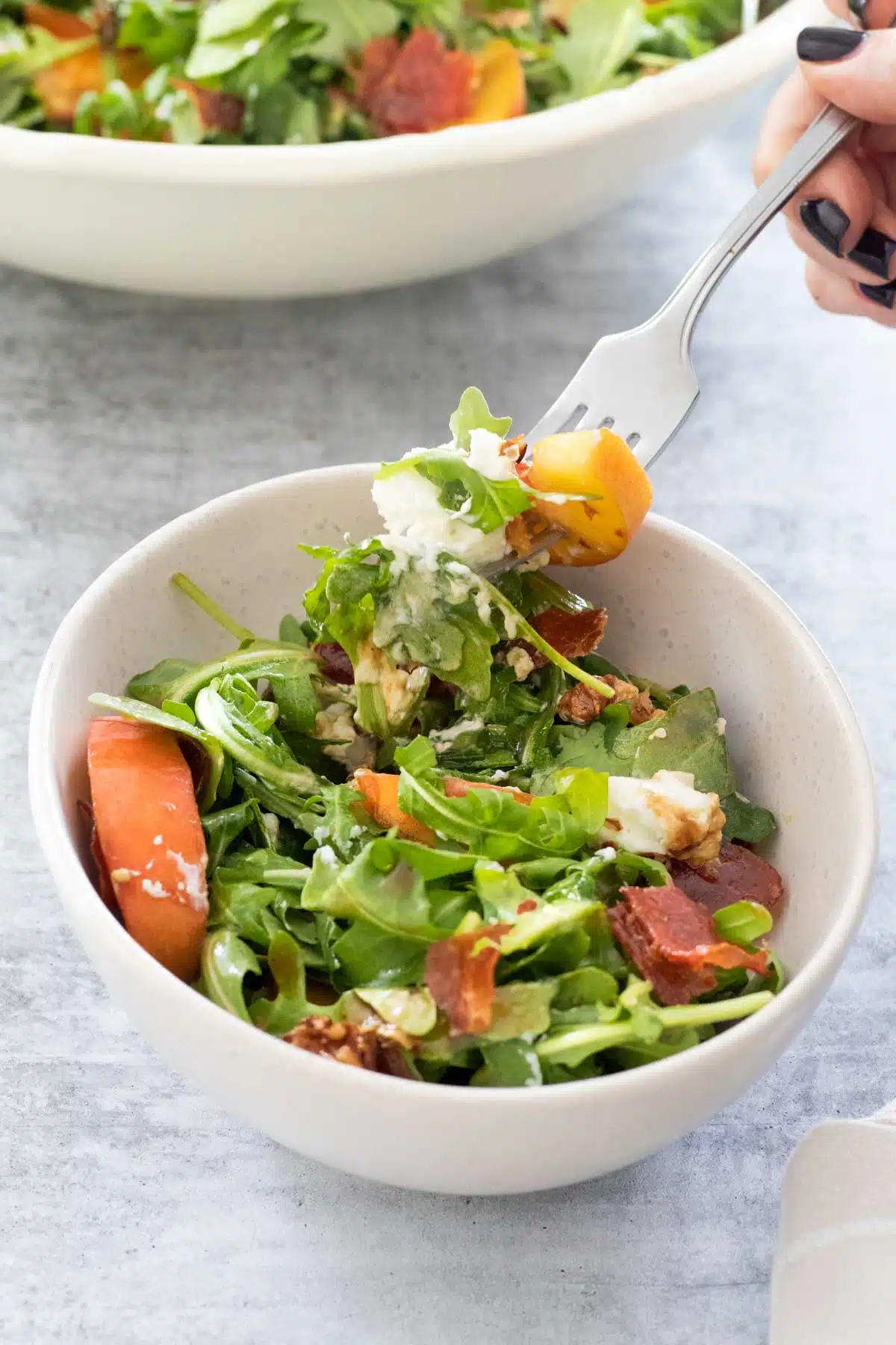 A white hand with black nail polish has a forkful of salad hovering above a small bowl. The larger bowl of peach arugula salad sits in the background.