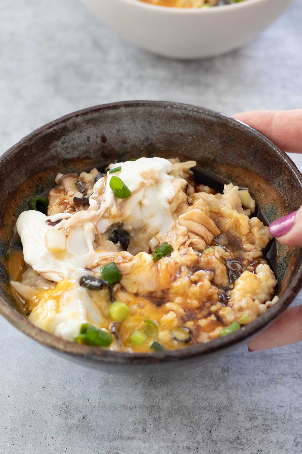 A brown bowl filled with savoury oats and a runny poached egg is being held by a white hand with dark pink nail polish.