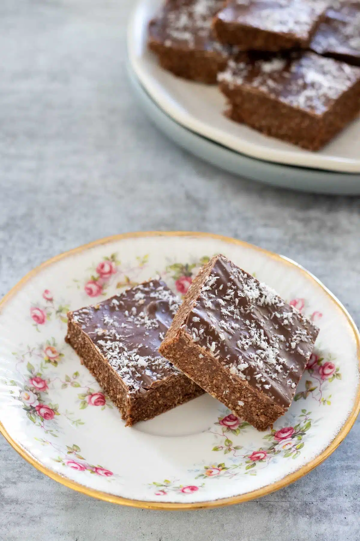 A small floral plate with two pieces of coconut chocolate slice is sitting on a grey table. A white plate stacked on a grey is at the top of the image, which is also filled with more slice.