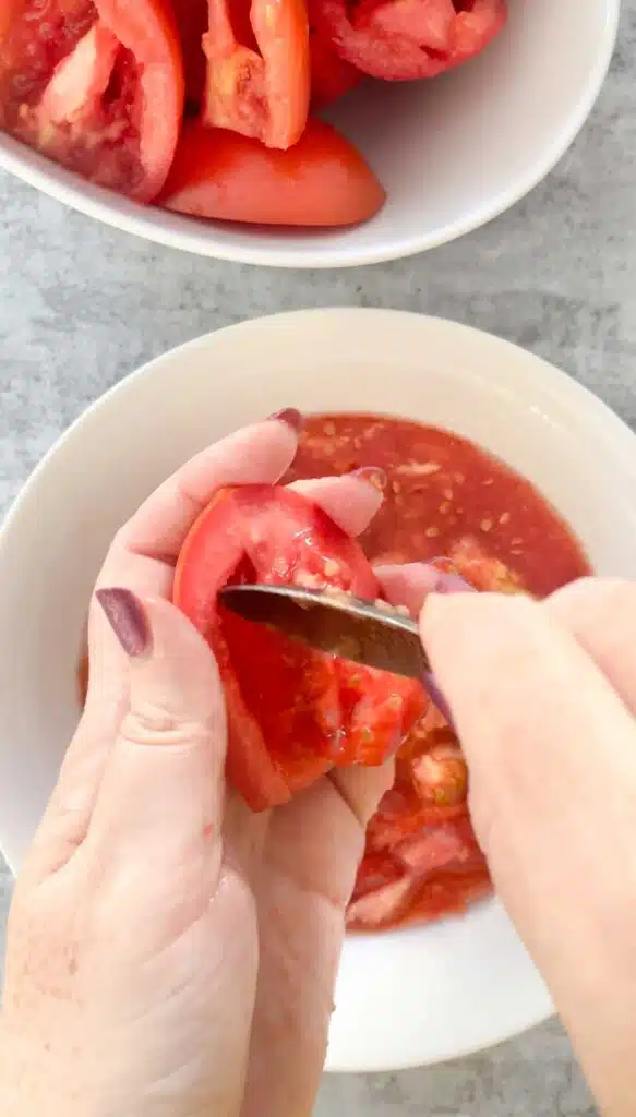 Two white hands with dark pink nail polish are de-seeding a tomato above a white bowl.