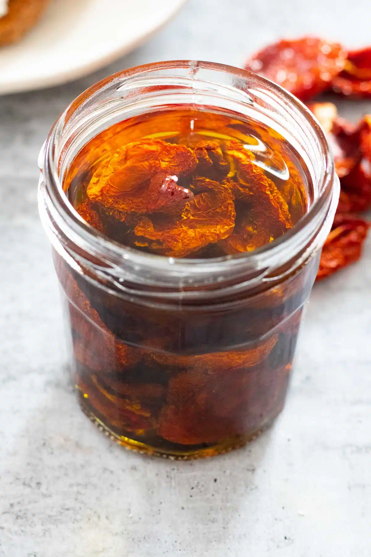 A jar of oven dried tomatoes has been covered in olive oil and is sitting on a grey table.