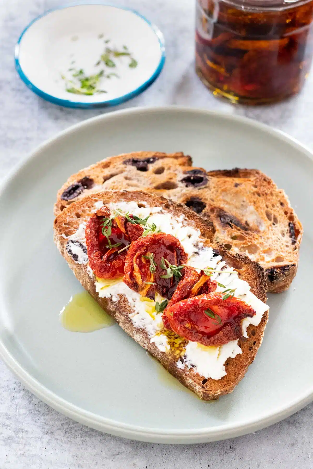 Two pieces of toast are on a duck egg green plate. One piece has been spread with goats cheese and topped with oven dried tomatoes drizzled with olive oil and sprinkled with fresh thyme. A jar of oven dried tomatoes in oil is just above the plate on the table.