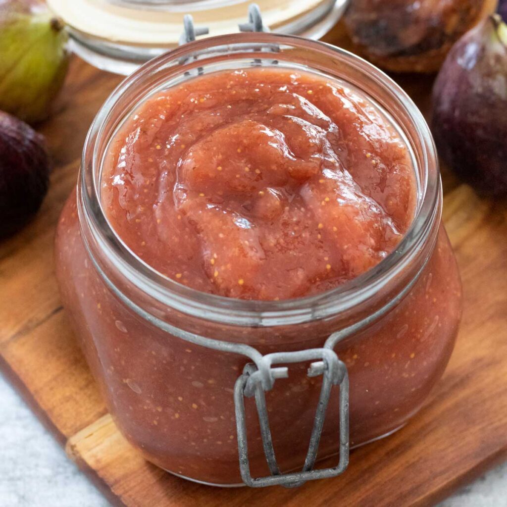 A jar full of fermented fig butter is sitting on a wooden serving board with figs and toast behind it.