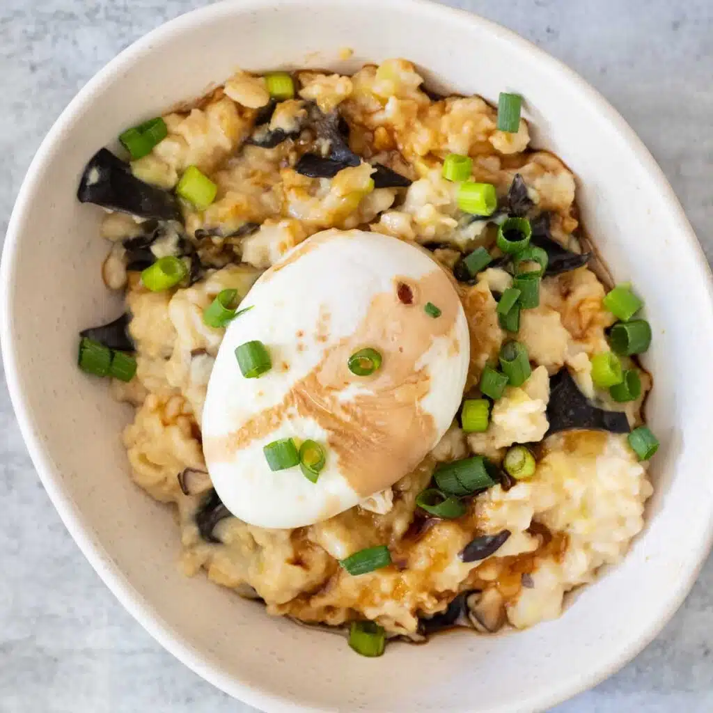 A square image showing a white bowl filled with savoury oats. Mushrooms, leek, and shallots are mixed through and a poached egg is sitting on top. Tamari and sesame oil have been drizzled over everything.