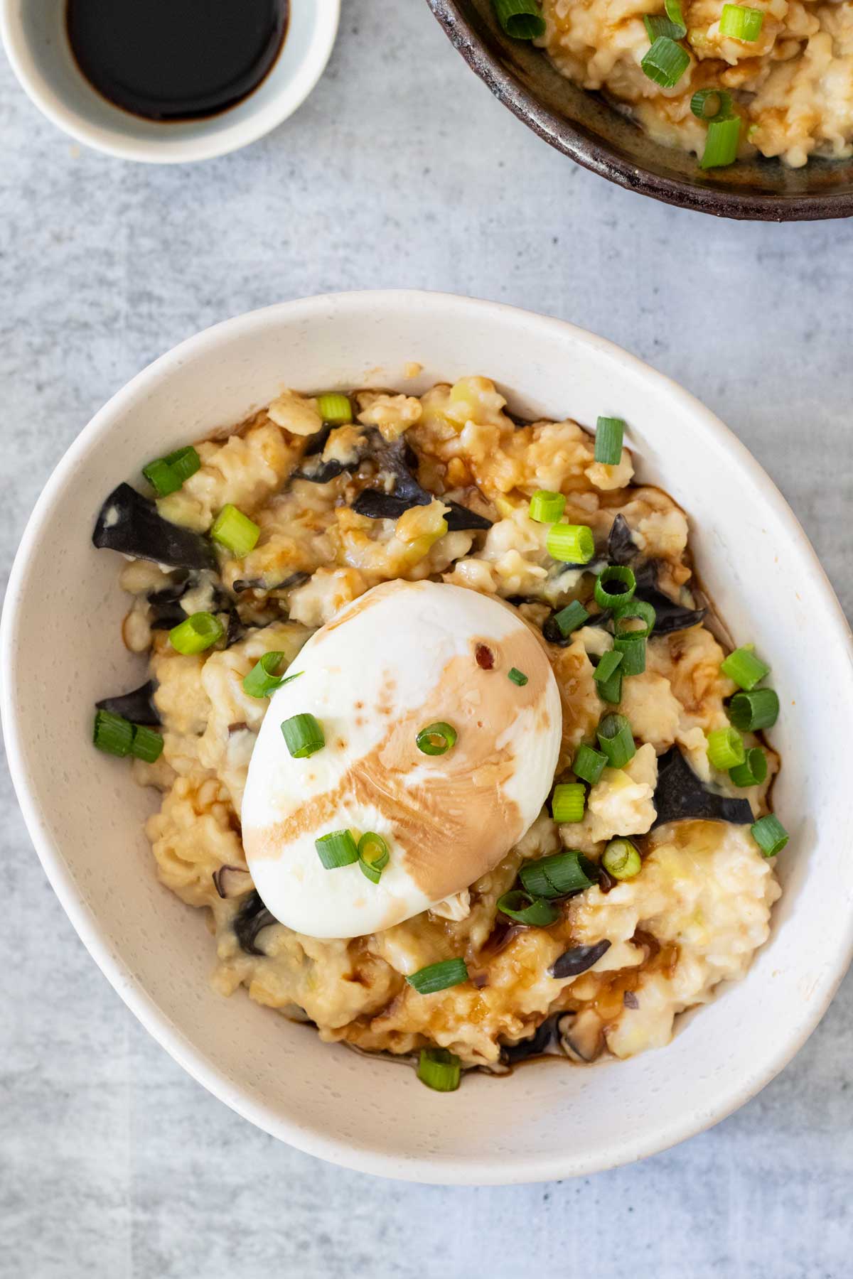 Overhead photo of a white bowl filled with savoury oats. Mushrooms, leek, and shallots are mixed in and a poached egg is sitting on the top. Tamari and sesame oil have been drizzled over everything.