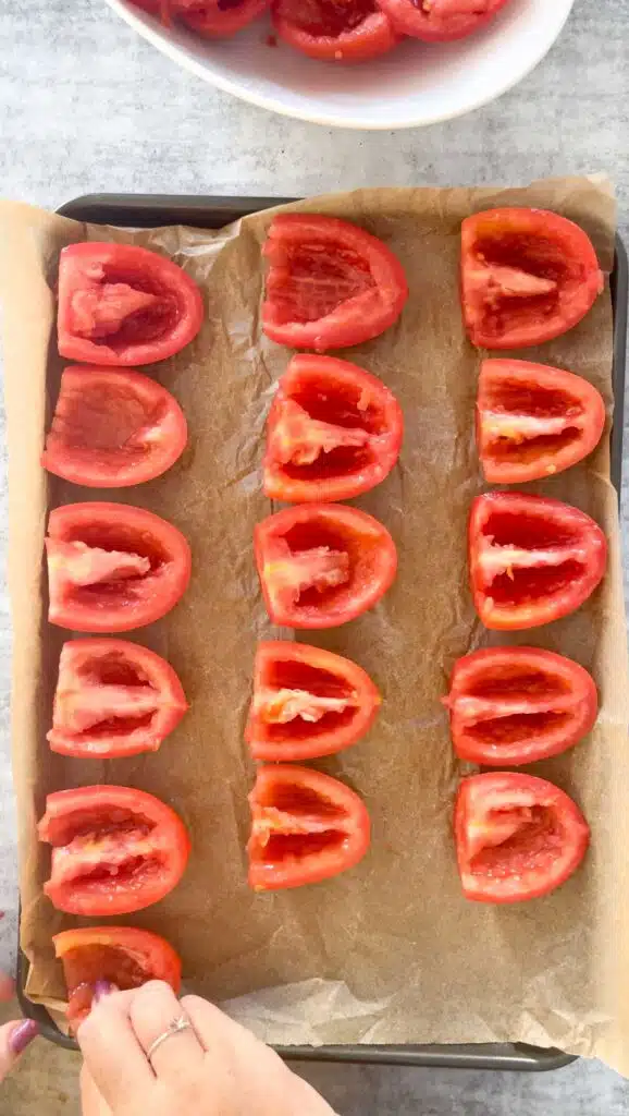 The de-seeded tomatoes are being placed cut side up on a baking tray lined with baking paper by two white hands that have dark pink nail polish.