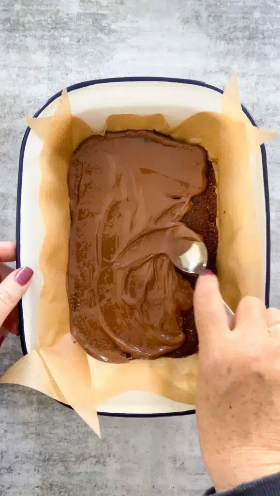 A white hand with dark pink nail polish is holding a white tin that contains the coconut chocolate slice. The other hand is smoothing over melted chocolate with a spoon for the topping.