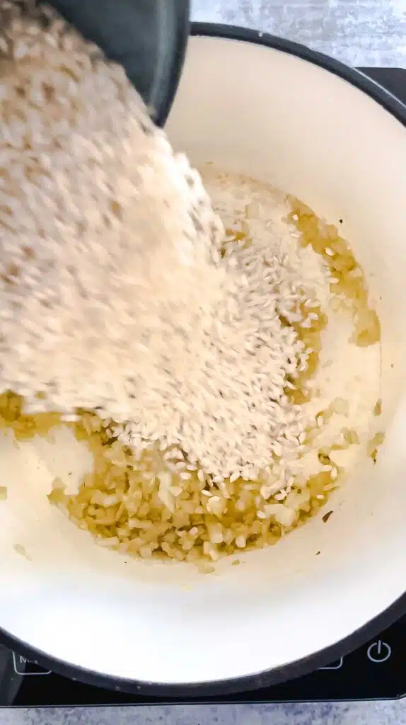 Rice is being poured into a heavy-based saucepan to be combined with cooked onion and garlic. The pot is sitting on a small induction cooktop.