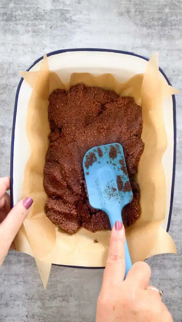 A white tin lined with baking paper contains the mixed ingredients for coconut chocolate slice. One white hand with dark pink nail polish is holding the tin. The other hand is pressing down the slice with a blue spatula.